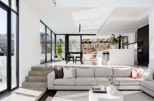 Bright open-plan contemporary interior combining a sunken living area with a large sectional sofa and a raised kitchen and dining zone. Full-height black-framed glazing brings in abundant daylight, while a white island, dark kitchen cabinetry, polished concrete or stone steps, and a planted stone retaining wall create a refined indoor-outdoor connection. A sculptural recessed ceiling detail and linear pendant add architectural interest.