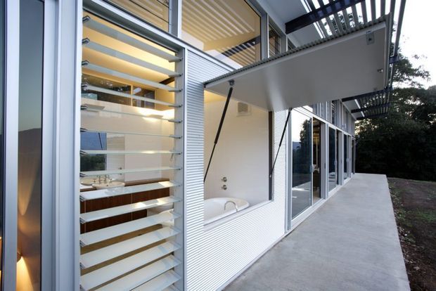 Exterior view of a contemporary multi-level home with extensive glazing, horizontal louvres, corrugated metal cladding, and a covered concrete terrace. A bathroom with a freestanding bath is visible through the open louvred opening, while the building facade and shaded outdoor circulation dominate the image.