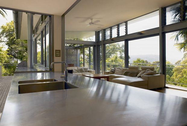 Open-plan kitchen and living area with a large stainless-steel island bench in the foreground, integrated double sink and mixer tap, and extensive floor-to-ceiling glazing. A neutral sectional sofa occupies the living zone, while a ceiling fan, dark window frames, and leafy elevated views create a bright indoor-outdoor connection.