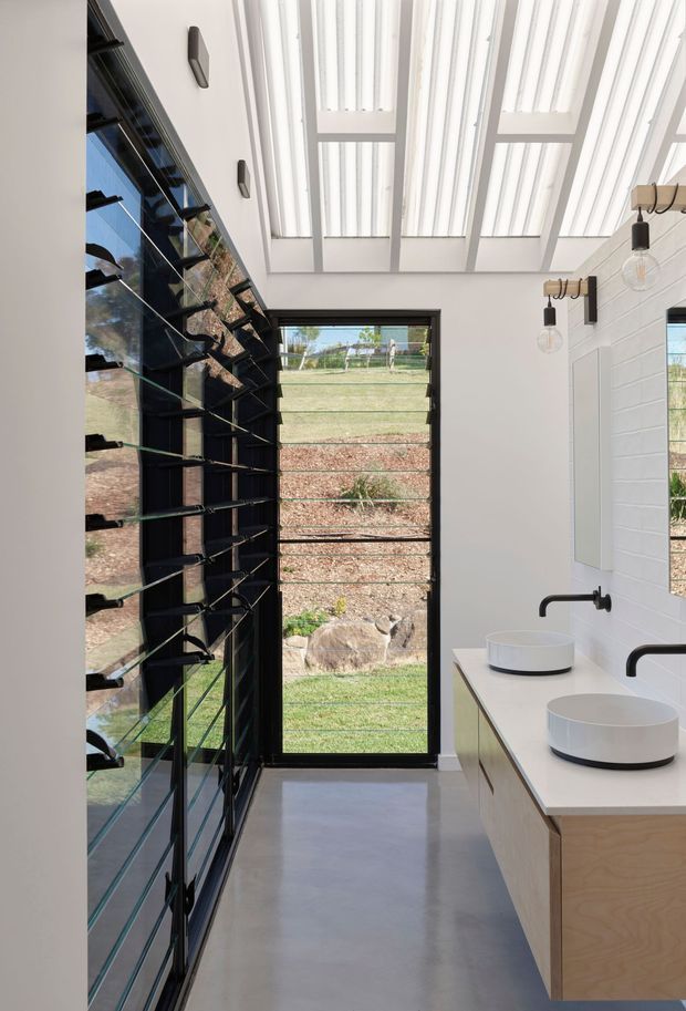 Bright contemporary bathroom with a long double vanity, two round vessel basins, wall-mounted black taps, white painted brick wall, pale timber vanity cabinetry, and a polished grey concrete floor. A dramatic black-framed glazed wine display wall lines the left side, while a large glazed door and extensive translucent roof panels bring in abundant natural light and views of the landscaped grounds.