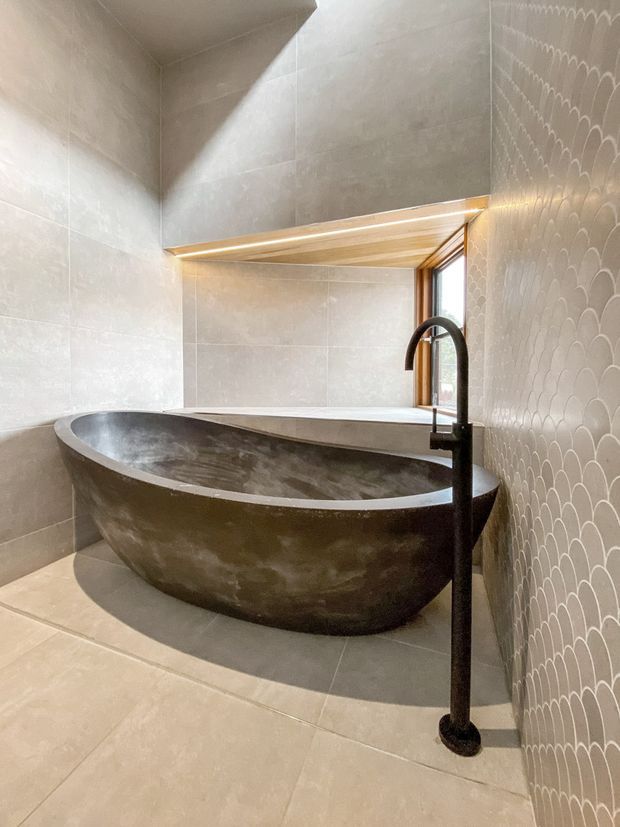 A refined bathroom featuring a freestanding dark stone or concrete bathtub, a tall matte-black floor-mounted faucet, large neutral wall and floor tiles, and a decorative scalloped tile feature wall. Warm concealed lighting highlights the timber-lined ceiling recess above the tub, with a window providing natural light.