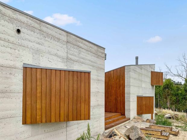 Exterior view of a minimalist contemporary building composed of pale board-formed concrete walls with warm vertical timber-clad shutters or panels. The structure has simple rectilinear volumes, a low-profile roofline, timber steps and decking, and a rugged landscaped site with exposed rocks and vegetation.