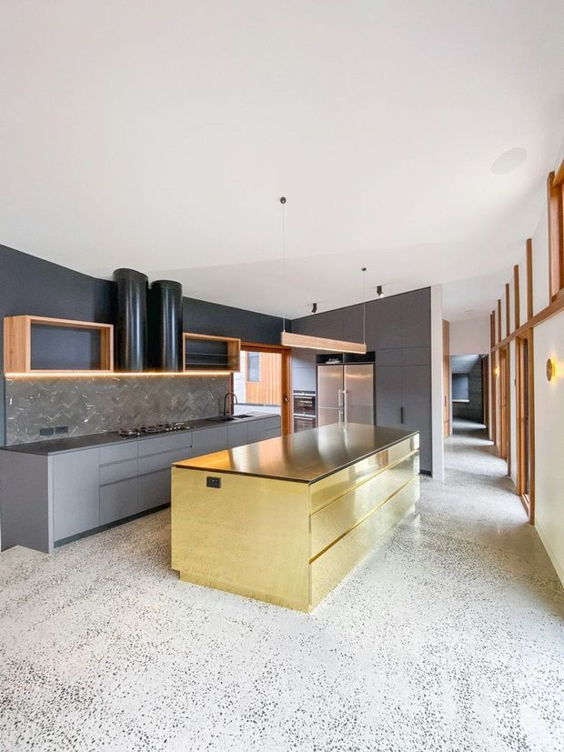 Bright contemporary kitchen with a large brass-toned island, dark grey cabinetry, terrazzo flooring, a patterned tiled backsplash, integrated appliances, and black cylindrical rangehoods. Timber-framed glazed doors and partitions line the right side, opening toward a hallway. The room has a clean, refined look with strong contrast between the dark joinery, metallic island, and pale floor and ceiling.
