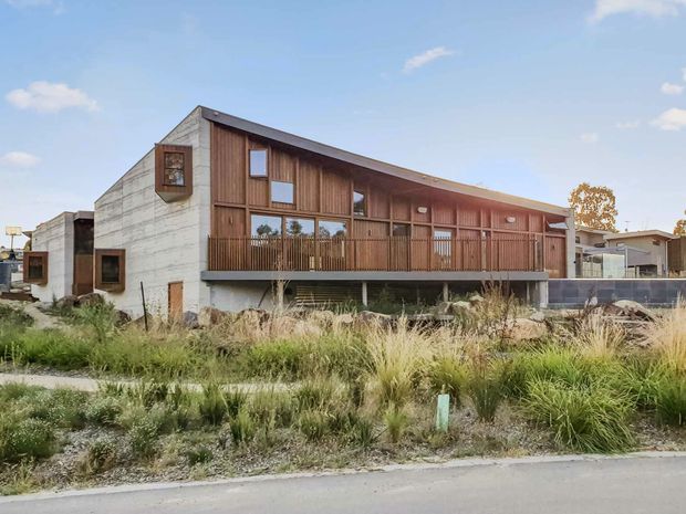 Exterior view of a contemporary two-storey building with a long, low-pitched roofline, exposed concrete side walls, and a warm timber-clad upper facade. A continuous timber balcony with vertical balustrading fronts large glazed openings, while landscaped grasses and a paved street occupy the foreground.