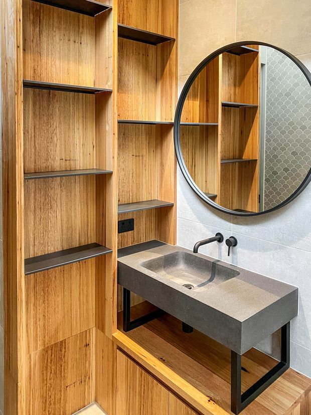 A contemporary bathroom vanity area featuring a custom timber wall of open shelving, a thick dark grey integrated basin benchtop, black tapware and supports, and a large round black-framed mirror. Light grey wall tiles and warm timber flooring create a refined, warm finish.