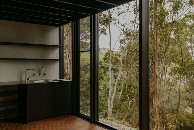 A dark, minimalist kitchenette or wet bar set against floor-to-ceiling black-framed glazing. The space features black built-in cabinetry, a slim countertop with sink and tapware, open shelving, a textured light wall finish, dark ceiling beams, and warm terracotta-toned tiled flooring. The glazing overlooks dense native woodland.