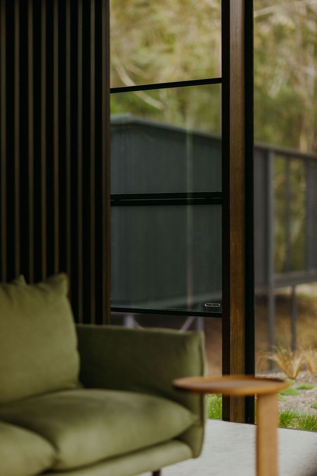 A moody interior detail showing a deep olive-green upholstered lounge chair beside a small timber side table. Full-height black-framed glazing and warm timber window framing look onto a wooded exterior and dark fence, while the interior wall features narrow vertical dark timber or black slatted detailing. The scene has a refined, organic character with soft natural light.