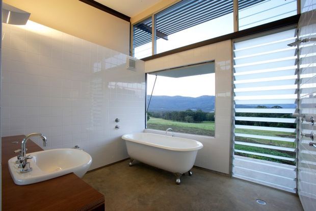 A bright contemporary bathroom with a freestanding white clawfoot-style bathtub, wall-mounted shower fixtures, a white basin on a timber vanity, glossy white tiled walls, and a polished concrete-look floor. Large windows and horizontal louvres provide views toward a green rural landscape and distant hills, while dark timber-framed glazing adds contrast.