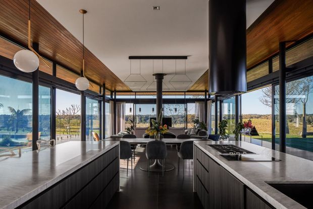A spacious contemporary kitchen and dining area with two long dark-wood islands, pale stone benchtops, integrated appliances, and sculptural pendant lighting. Full-height glazing wraps the room, opening views to the landscaped grounds and distant rural surroundings. A dramatic black cylindrical rangehood forms a central feature, while timber-lined ceiling and wall sections add warmth to the otherwise refined, open-plan interior.