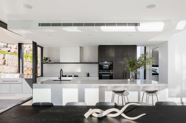 Bright, open-plan contemporary kitchen and dining area with a large white island, integrated appliances, dark timber cabinetry, pale benchtops, and a long dark dining table in the foreground. Full-height glazing and a stone retaining wall are visible to the left, bringing in abundant natural light. Minimal decor, clean linear detailing, and indoor plants create a refined modern interior.
