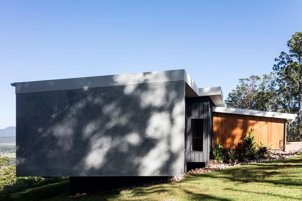 Exterior view of a compact contemporary house on a grassy, gently sloping site. The building has strong rectilinear volumes, a dark smooth rendered facade, black vertical entry glazing and cladding, and a warm timber-clad wing. Trees and distant hills form the background.