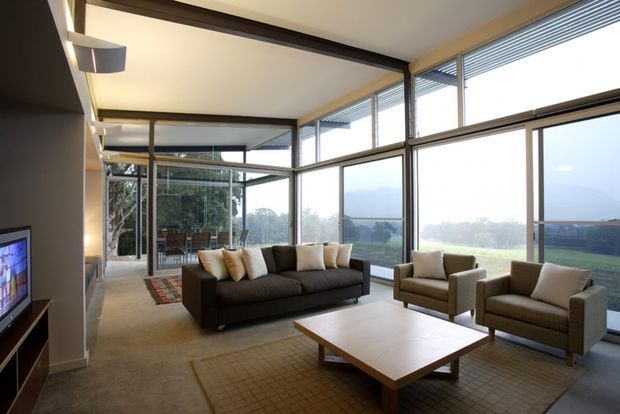 A spacious contemporary living room with expansive floor-to-ceiling glazing on two sides, offering views of a green landscape. The room has a dark sectional sofa, two upholstered armchairs, a large low timber coffee table, area rugs, recessed linear lighting, and integrated media cabinetry. The glazing includes horizontal blinds and dark slim frames, creating a bright, open connection to the outdoors.