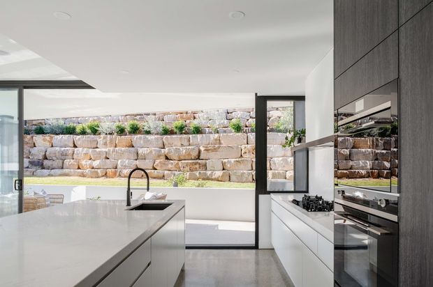 Bright contemporary kitchen with a large white island and integrated sink, white handleless cabinetry, dark wood-grain tall cabinetry, built-in ovens, and a gas cooktop. Full-height glazing opens views toward a landscaped stone retaining wall, with polished concrete flooring and a clean minimalist fit-out.