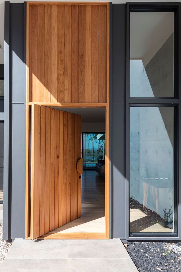 A modern residential entrance viewed from outside, featuring a tall pivot-style timber door with vertical slat detailing, set within a dark charcoal facade. Full-height black-framed glazing sits beside the entry, reflecting and revealing concrete surfaces and glimpses of the interior beyond. A concrete path leads to the doorway, with dark gravel landscaping at the base.