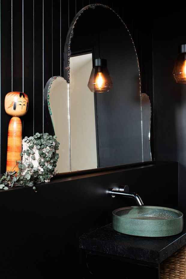 A moody, dark bathroom vanity vignette with black vertically panelled walls, a large irregular-edge arched mirror, warm pendant lighting, and a green textured vessel basin on a dark stone benchtop. A timber decorative figure and trailing plant sit beside the mirror.