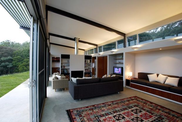 A spacious contemporary living room with a double-height ceiling and a mezzanine-like upper band of clerestory windows. Full-height glazing and an open sliding door connect the room to a grassy, wooded exterior. The room features built-in shelving around a television, a long built-in window seat, dark sofas, a patterned area rug, recessed lighting, and exposed dark structural framing.