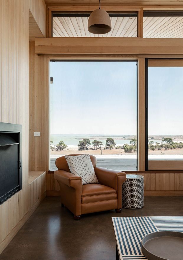 A warm, timber-lined living room with large black-framed windows overlooking a coastal or lakeside landscape. The room features a caramel leather armchair, patterned side table, striped rug, round coffee table, pendant light, built-in timber cabinetry, and a partially visible television and fireplace surround. The interior has polished concrete flooring and extensive natural light.