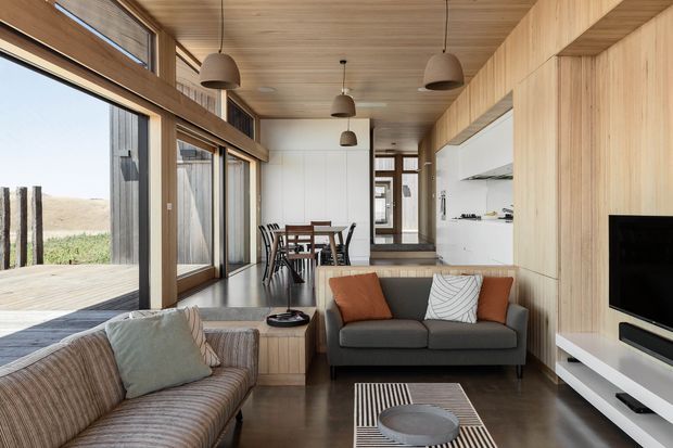 A warm, compact open-plan living room with extensive timber wall and ceiling lining, full-height glazing opening to a timber deck, a grey sofa and striped armchair, and a dining area positioned toward the rear. Built-in timber cabinetry houses a wall-mounted television, while pendant lights and a restrained neutral palette create a refined contemporary retreat.