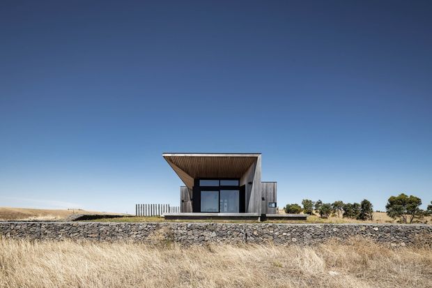 A minimalist single-storey modern pavilion-like building set in a dry rural landscape. The structure features a strong cantilevered flat roof, exposed timber soffit, dark-framed full-height glazing, and weathered grey exterior walls, with a low stone retaining wall across the foreground.