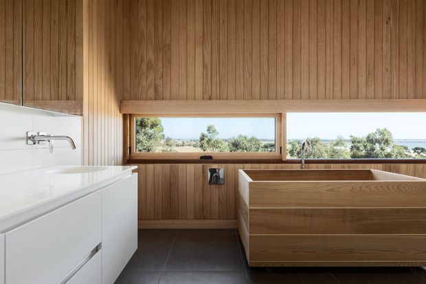 A contemporary bathroom with a freestanding timber bathtub, white wall-mounted vanity, brushed metal tapware, dark grey floor tiles, and extensive vertical timber wall panelling. A wide horizontal window provides views toward greenery and water beyond.