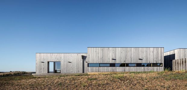 A low, elongated contemporary building in a rural open setting, clad in weathered vertical timber boards. The simple composition includes a taller central volume, a lower entrance wing, and narrow horizontal strip windows beneath a clear blue sky.