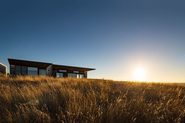 A low, modern single-storey house with dark cladding and extensive glazing sits within tall golden grasses. The flat-roofed structure is viewed from the surrounding landscape at sunset, with strong horizontal lines and a broad overhanging roof.