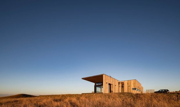 A modern timber-clad house set prominently in a broad, open grassy landscape. The low, angular building features projecting roof planes, extensive glazing, and a simple contemporary composition beneath a clear blue sky.