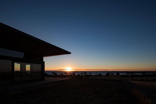 Dark dusk exterior of a modern single-storey building with a broad projecting roof and glazed wall, silhouetted against a low sunset over a coastal or waterfront horizon. The surrounding site is mostly dark and indistinct.