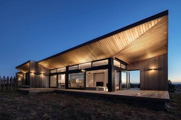 Contemporary single-storey house photographed at dusk, featuring a dramatic asymmetrical sloping roof with a deep timber-lined soffit, dark vertical timber cladding, extensive black-framed glazing, and a timber deck. Warm interior lighting reveals an open-plan living area through the glass, while the building sits in a sparse coastal or rural setting.