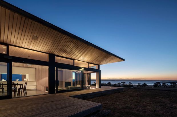 Twilight exterior view of a modern single-storey home with broad timber-lined roof eaves, dark framed floor-to-ceiling glazing, and a timber deck overlooking a calm coastal or waterfront landscape. The open-plan kitchen and living area are visible through the glazing, with warm interior lighting contrasting against the blue evening sky.
