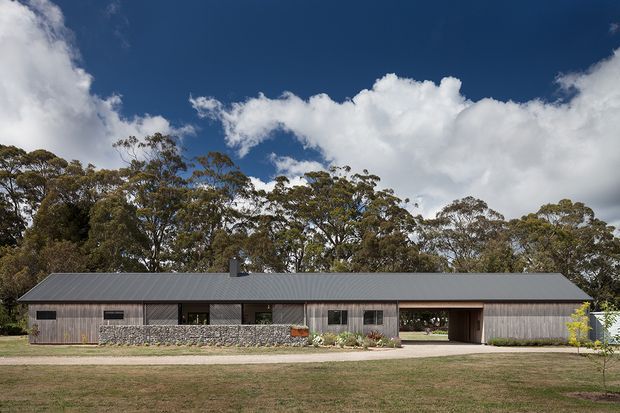 A long, low single-storey rural residence set in a landscaped bush setting. The building has a simple contemporary linear form, a dark pitched metal roof, weatherboard-style vertical cladding, extensive glazing, and a low stone retaining or boundary wall. A covered vehicle passage or carport divides the main facade, with mature eucalyptus trees and open lawn surrounding the house.