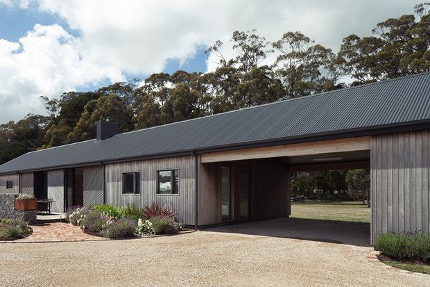 Single-storey rural-style building viewed from the front, with a long dark corrugated metal pitched roof, vertical timber-clad walls, a covered vehicle passage through the building, and small windows and doors. A gravel forecourt, brick-paved entry path, landscaped garden beds, and mature trees surround the structure.