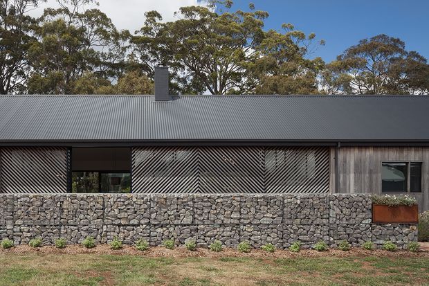 Single-storey contemporary residential facade set behind a low gabion stone wall. The building has a long dark corrugated metal pitched roof, weathered timber cladding, and distinctive diagonal timber screening across the glazing. Native trees and a simple lawn surround the structure, creating a restrained, natural Australian bush setting.