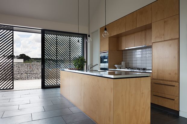 A bright contemporary kitchen with extensive warm timber cabinetry, a large central island with a dark benchtop, integrated appliances, and a white rectangular-tile splashback. Grey stone-look floor tiles extend toward full-height glazed doors with diagonal screening, opening to an exterior stone wall. Pendant lights hang above the island, and a small indoor plant adds greenery.
