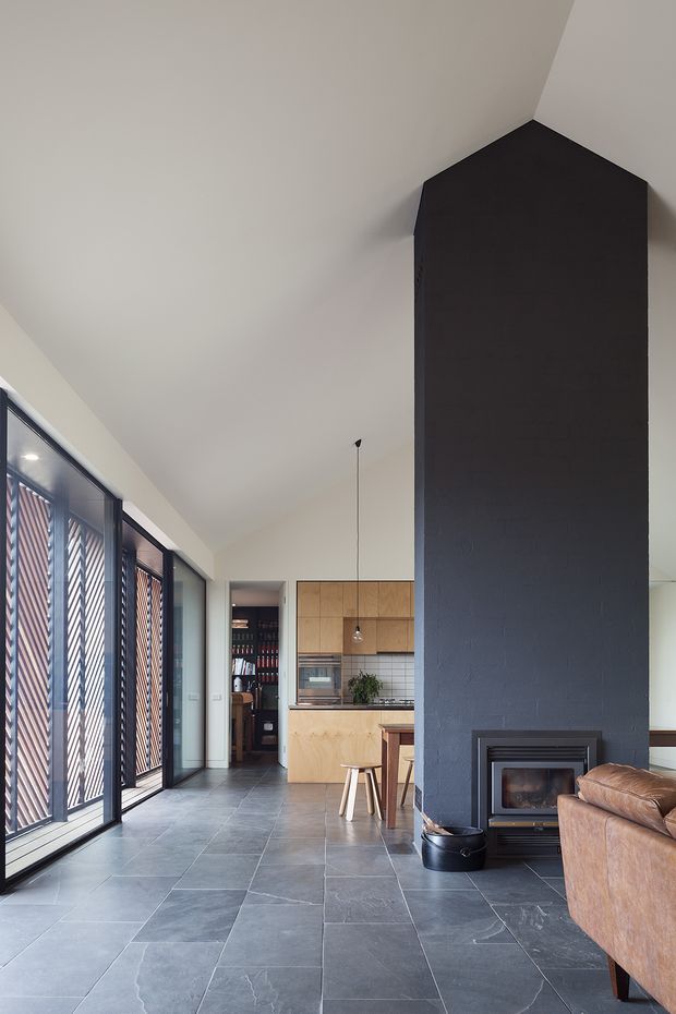 A contemporary open-plan living and kitchen interior with a tall dark masonry fireplace chimney forming a strong central architectural feature. The room has a high sloping ceiling, large glazed doors with patterned external screening along the left side, dark slate-like floor tiles, and warm timber kitchen cabinetry at the rear. A built-in wood-burning fireplace, leather sofa, kitchen island or peninsula, stools, and a pantry or storage area are visible.