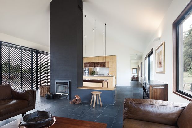 A bright contemporary open-plan living space with a central dark tiled fireplace and chimney mass, integrated kitchen behind it, and large windows along one side. Brown leather sofas and a timber coffee table furnish the lounge, while slim pendant lights hang above the kitchen island. A black diagonal-slatted screen forms the backdrop to the fireplace, with framed artwork and timber cabinetry along the window wall.