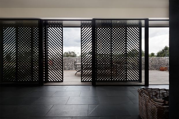 A dark, refined interior opening onto an outdoor terrace through large glazed openings fitted with black timber lattice screens. The room has large dark rectangular floor tiles and a woven armchair in the foreground. Outside, a pale stone wall, timber decking and greenery are visible.