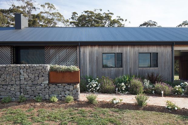 Single-storey contemporary house facade with a long corrugated metal gable roof, weathered vertical timber cladding, dark-framed windows, and a covered entry at the right. A gabion stone wall with a rusted corten-steel planter fronts the home, with a paved path and landscaped garden beds. Native trees form the background.