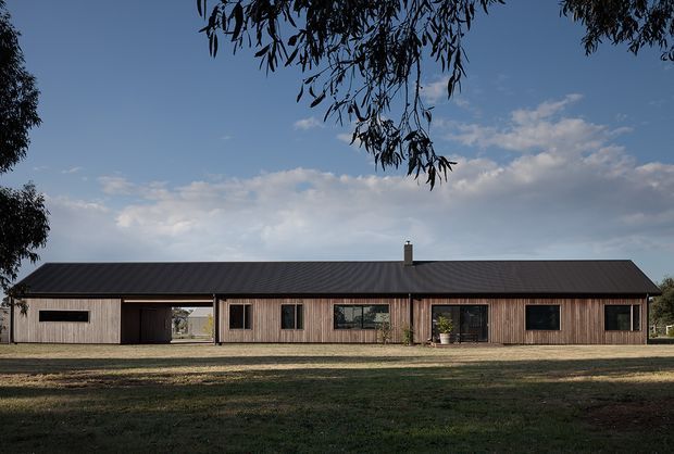 A long, low single-storey rural residence viewed straight on from the front. The house has a dark pitched metal roof, extensive vertical timber cladding, dark-framed rectangular windows, and a covered recessed entry or breezeway near the left side. It sits in an open grassy landscape beneath mature trees and a partly cloudy sky.
