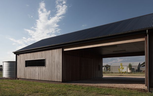 Exterior view of a simple rural outbuilding or barn-like structure with vertically clad timber walls, a dark corrugated pitched roof, and a large open-sided covered bay. A small horizontal window and cylindrical water tank are visible, with additional agricultural buildings seen beyond the opening.