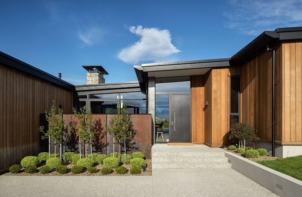 Contemporary single-storey residential exterior with a sheltered entry courtyard. The building combines warm vertical timber cladding, dark metal roof and window frames, expansive glazing, and a concrete or light aggregate-paved approach. Carefully clipped shrubs and young trees line the entry, while a distant landscape is visible through the central glazing. The composition features strong horizontal roof planes and a clean, modern material palette under a bright blue sky.