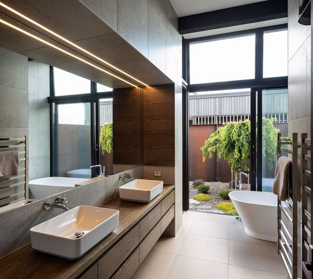 Contemporary bathroom with a double vanity, large mirror, freestanding bathtub, and floor-to-ceiling black-framed glazing opening onto a landscaped courtyard. The room features large grey wall and floor tiles, dark timber-look vanity cabinetry, a warm wood feature wall, integrated linear ceiling lighting, and a towel warmer. The bathroom has a spacious, bright, refined character with strong indoor-outdoor connection.