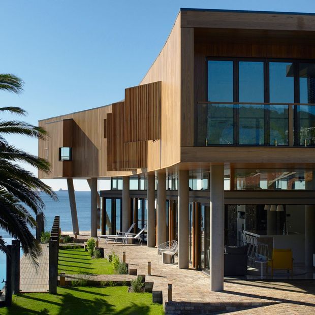 A contemporary multi-level coastal residence overlooking the ocean, with extensive glazing, projecting upper volumes, vertical timber cladding, covered outdoor living areas, stone-paved terraces, and landscaped lawn with palm trees. The open ground level reveals a kitchen or living area, while the main visual focus is the building exterior.