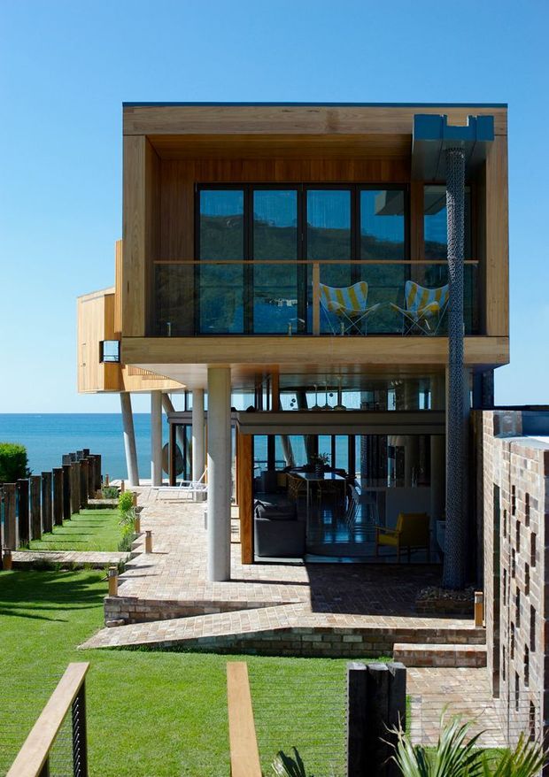 A contemporary multi-level coastal house viewed from the exterior, with a cantilevered upper volume clad in warm timber, extensive dark-framed glazing, and a covered ground-floor living area opening onto paved terraces and lawn. The house overlooks the ocean, with outdoor chairs visible on the upper balcony and a brick boundary wall to the right.