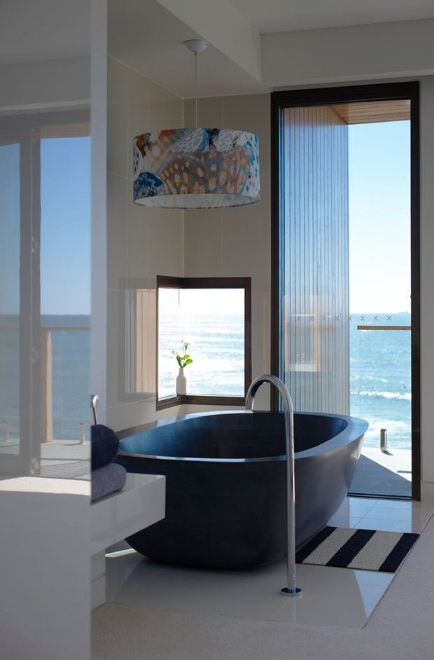 A bright, luxurious bathroom with a freestanding dark blue-black bathtub positioned beside full-height ocean-facing glazing and a smaller window. A tall chrome floor-mounted bath filler stands beside the tub, with pale tiled walls and floor, a patterned pendant light, and a striped bath mat. The ocean view is visible through the glazing, creating a coastal atmosphere.