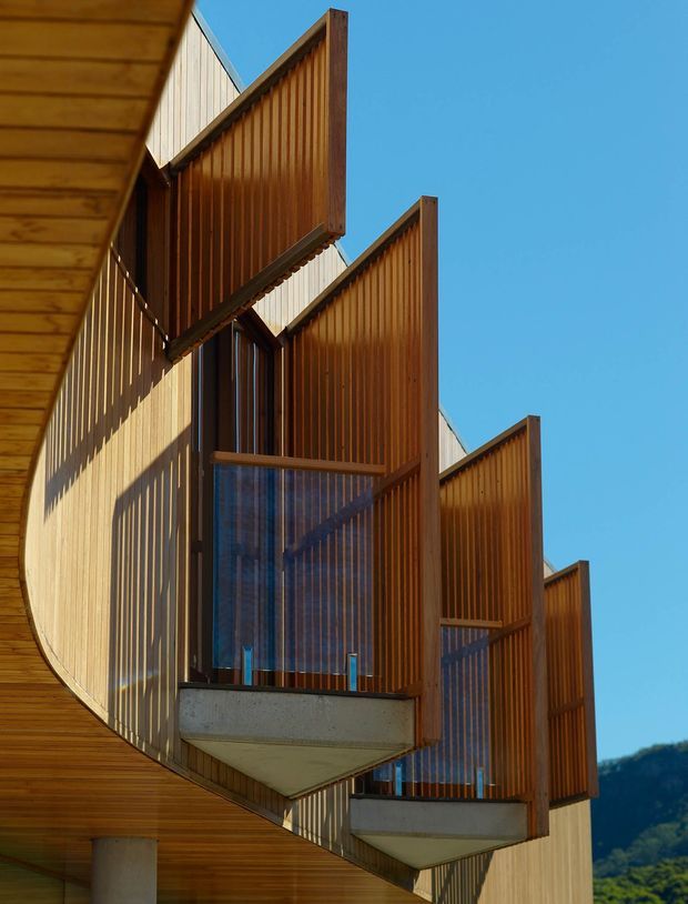 Upward-looking architectural exterior view of a contemporary multi-level building with projecting balconies, timber-clad soffits, vertical timber privacy screens, and translucent or glazed balustrades. Strong sunlight creates repeating linear shadows across the warm timber facade. A clear blue sky and distant wooded hillside are visible in the background.
