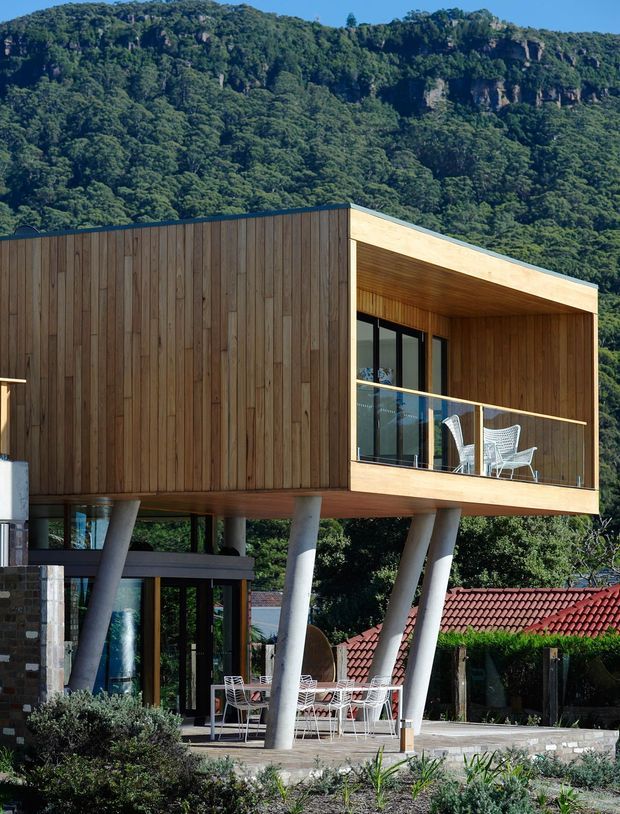 Exterior view of a contemporary elevated house set against a densely forested, rocky hillside. The upper level is clad in warm vertical timber boards and forms a deep covered balcony with timber soffit, glass balustrade and outdoor chairs. The lower level is supported by angled concrete columns and includes extensive glazing, a stone wall and an outdoor dining area. A neighbouring red tiled roof is visible in the background.