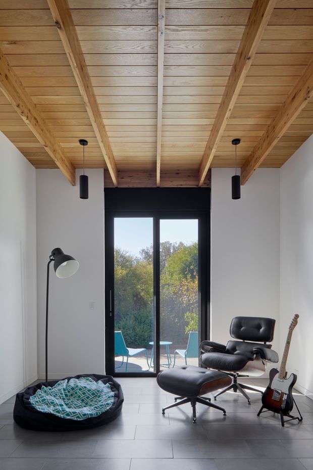 A compact, bright sitting or music room with white walls, large black-framed glazed doors opening to an outdoor area, and a distinctive exposed timber-beam ceiling with timber boards. The room contains a black leather lounge chair and ottoman, a floor lamp, a guitar, and a pet bed. Large-format pale grey floor tiles and black pendant lights create a clean, contemporary look, while the timber ceiling adds warmth.