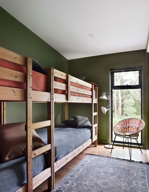 A compact children's bedroom with two natural timber bunk beds, dark green walls, and layered bedding in earthy red, brown, blue-grey, and charcoal tones. A black-framed window provides a view of trees, while a woven accent chair, slim floor lamp, patterned rug, and light timber floor create a warm, rustic and slightly eclectic atmosphere.