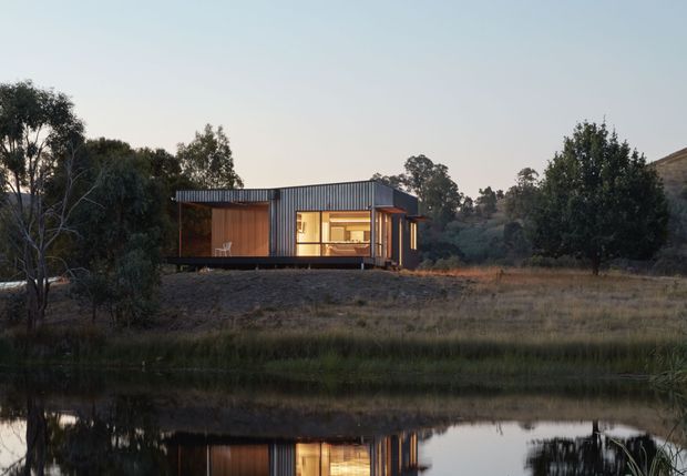 A compact modern single-storey dwelling set in a rural, lightly wooded landscape beside a reflective pond. The building has a simple flat-roofed rectangular form, dark corrugated metal exterior cladding, extensive glazing, and warm timber-lined interior or sheltered outdoor areas illuminated at dusk. The low-profile structure is surrounded by native vegetation and dry grassland.